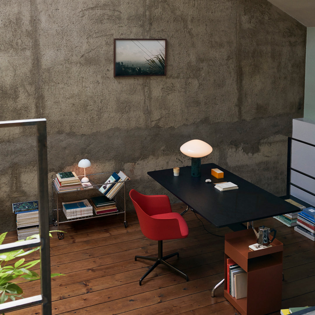 Modern workspace featuring a red chair, black desk, and earthy tones with a concrete wall backdrop. Chic decor accents.