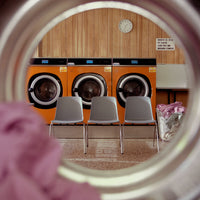 "Bright orange washing machines with gray chairs and a wooden backdrop, featuring a soft purple fabric in the foreground."