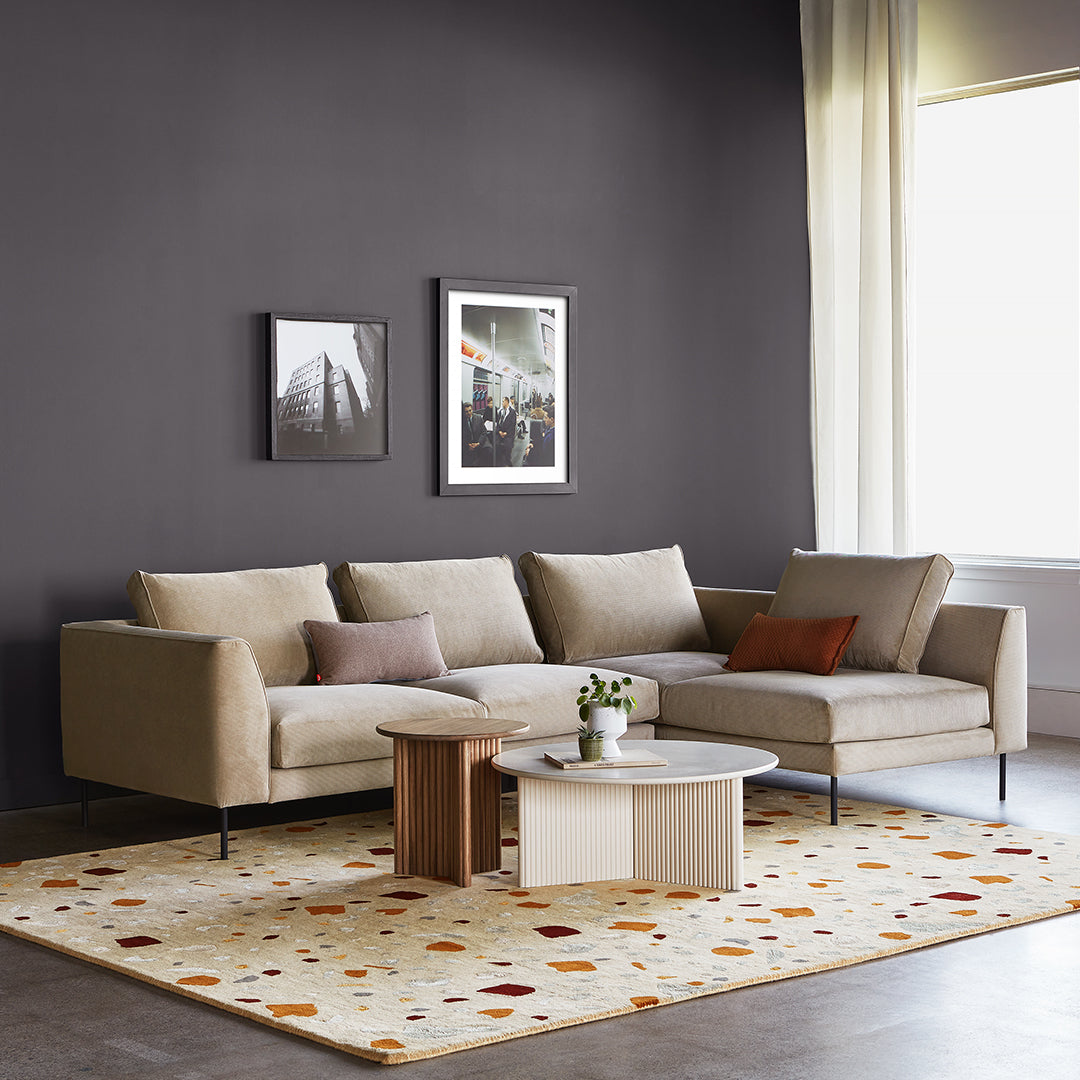 Beige sectional sofa on a light, speckled pattern rug with warm orange and earth tones, set against a dark gray wall.