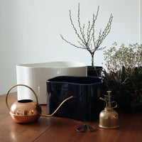 Modern planters in glossy white and deep navy, complemented by copper and brass watering can, surrounded by greenery.