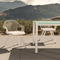 White wire outdoor chairs with soft cream cushions beside a sleek glass-top table in a sunny landscape.