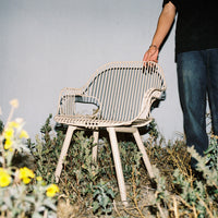 Light beige chair with vertical stripe pattern, set against a natural outdoor backdrop with green vegetation and yellow flowers.