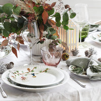 Elegant table setting featuring white plates with floral patterns, green foliage, and soft pink glassware against a neutral backdrop.