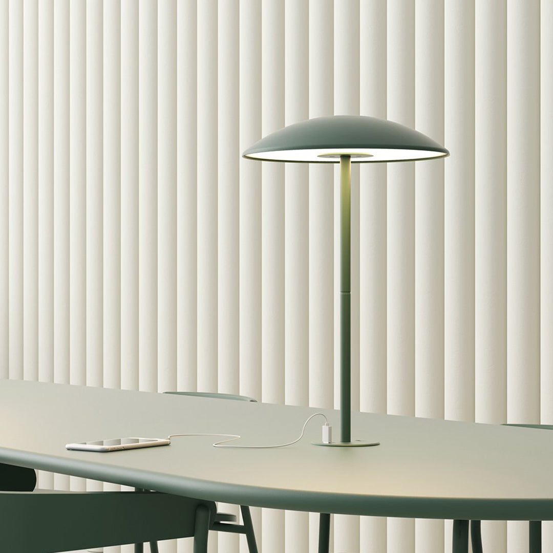 Modern green table lamp with a sleek dome shade, standing on a minimalist light-colored table against vertical cream blinds.