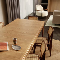 Natural wood dining table with light oak finish, complemented by woven cane chairs and soft neutral textiles in a modern setting.