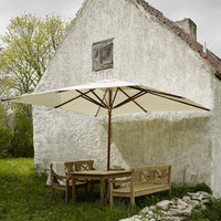 Cream-colored patio umbrella with a simple design, set against a rustic white wall and green grass backdrop.