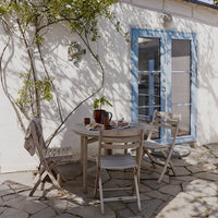 "Light wood outdoor furniture with neutral-colored cushions against a white wall and blue door."