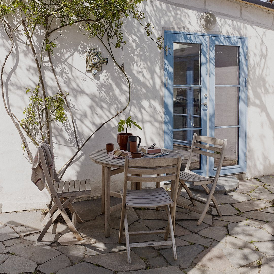 Quaint outdoor dining set featuring muted wood tones and a soft blue door, surrounded by natural greenery and stone surface.