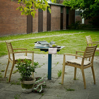 Wooden patio chairs in soft beige with a natural finish, set against a lush green lawn and rustic brick backdrop.