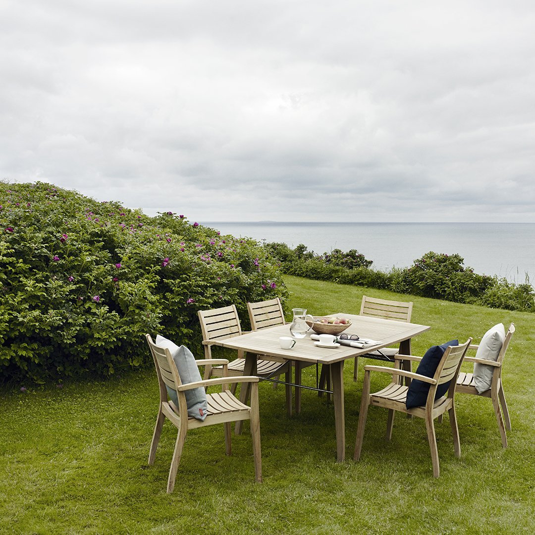 Light wood dining set with soft blue and charcoal cushions, surrounded by lush green grass and a cloudy sea backdrop.
