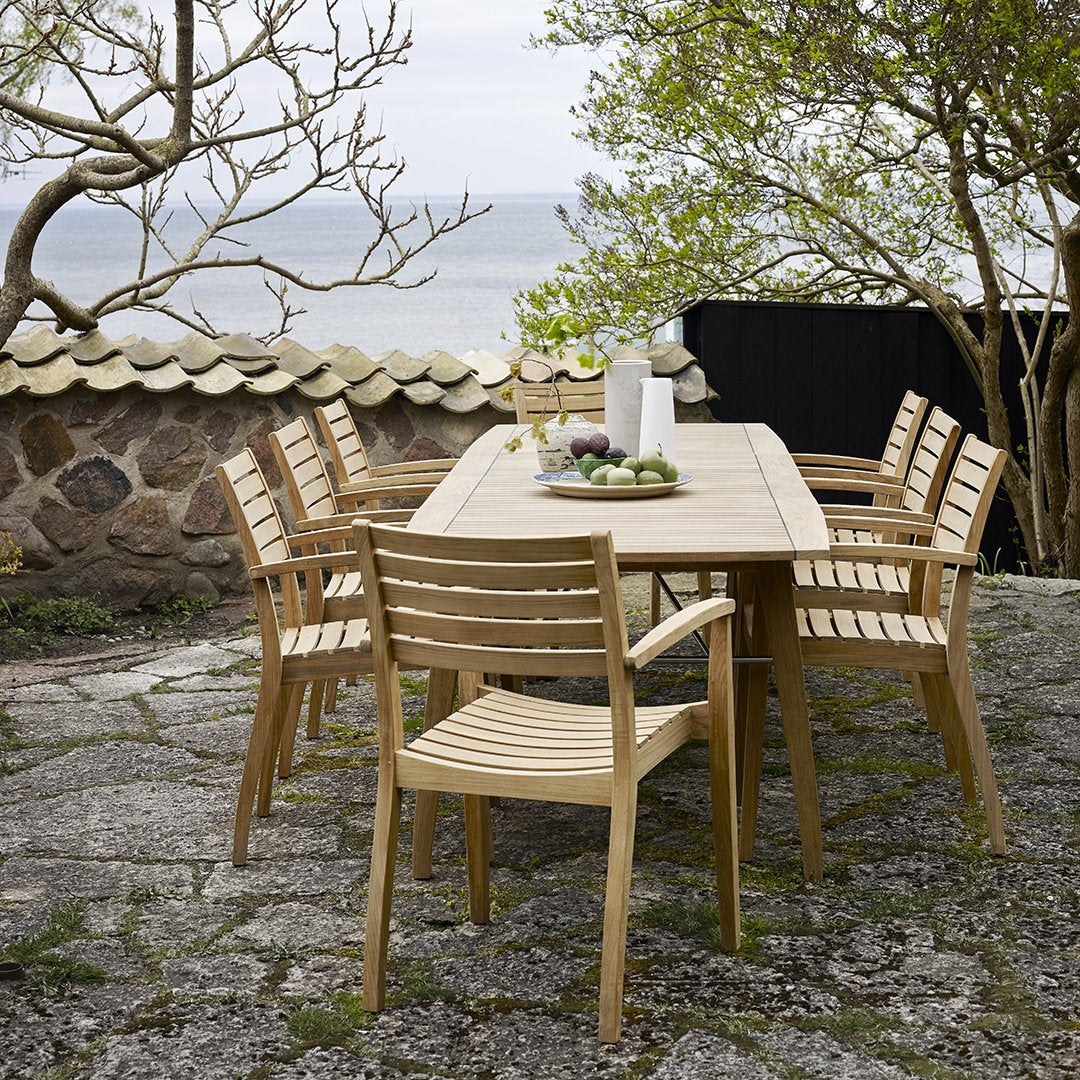"Natural wood outdoor dining set with a light finish and striped chair backs, set against a scenic coastal backdrop."