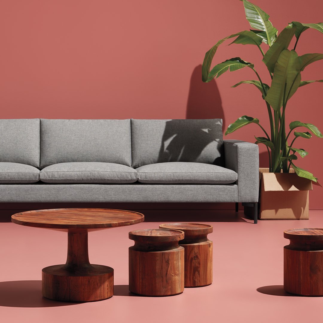 Gray sofa on a soft pink floor, with wooden circular tables and green foliage against a warm coral wall.
