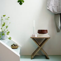 Wooden side table with natural finish, white candle, and brown bowl, set against a soft green and white backdrop.