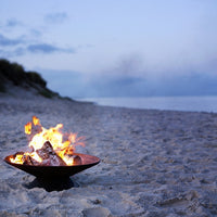 Copper fire pit with glowing orange flames on a sandy beach at dusk, complemented by soft blue-gray sky hues.