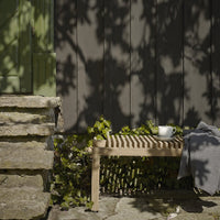 Light wood bench with slatted design against a dark gray wall, surrounded by green foliage and a gray throw.