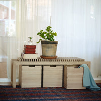 "Light wood storage bench with natural slats, complemented by a green plant and a teal blanket on a blue-patterned rug."