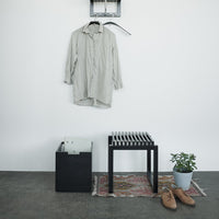 Light gray shirt hanging against a white wall, paired with a dark wooden stool, patterned rug, and green plant.