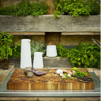 Wooden chopping board with vibrant green herbs, white kitchenware, and earthy vegetables in a rustic setting.