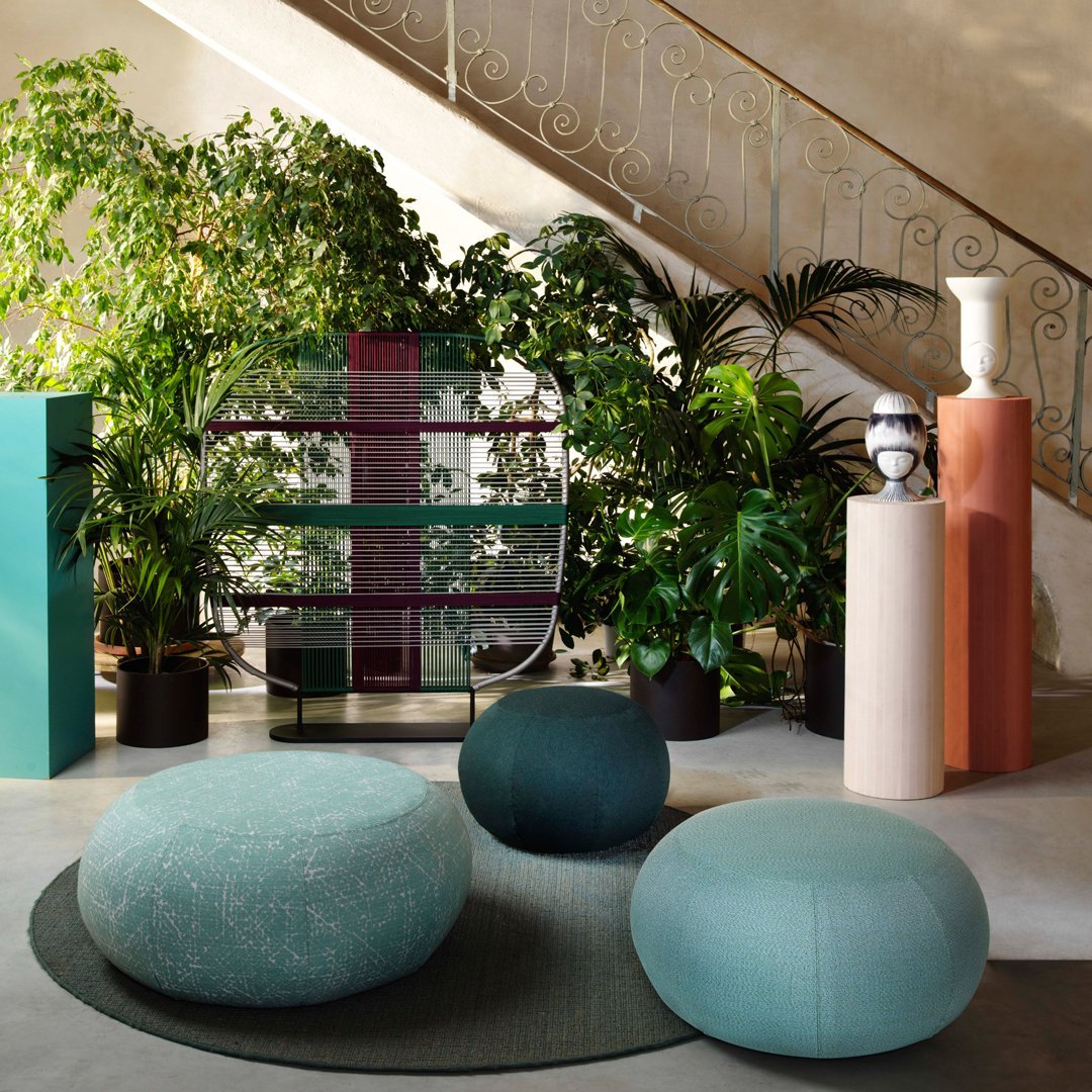 Turquoise and teal poufs on a textured rug, surrounded by green plants, neutral-colored sculptures, and colorful vertical storage.