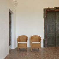 Pair of woven rattan chairs with soft cream cushions, set against a neutral wall and rustic wooden door.