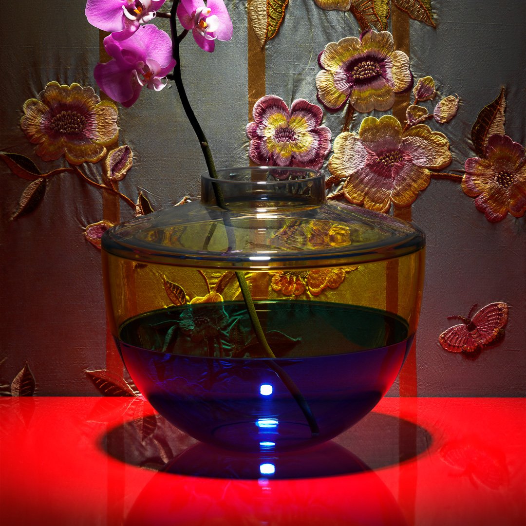 Colorful glass vase with yellow and blue layers, illuminated by LED lights, featuring a pink orchid and floral background.
