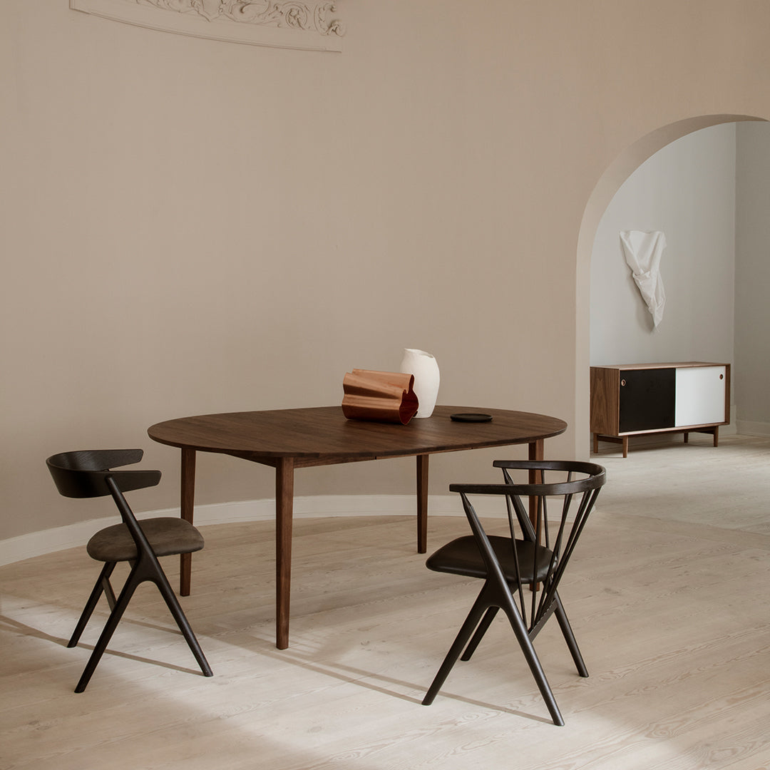 Modern wooden dining table with dark wood and black chairs, set against a neutral beige wall and light wooden flooring.