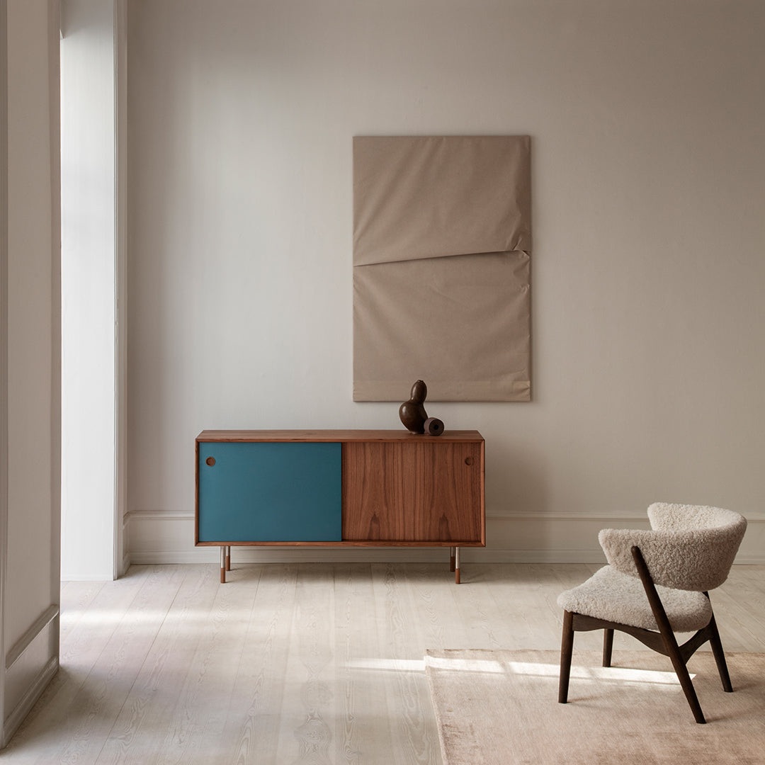 Wooden sideboard with a teal door, beige fabric wall art, and neutral tones in a minimalist interior.
