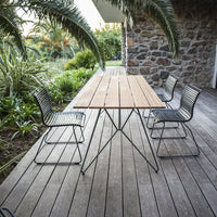 Outdoor dining set featuring a natural wood tabletop and sleek black metal chairs against a stone backdrop.