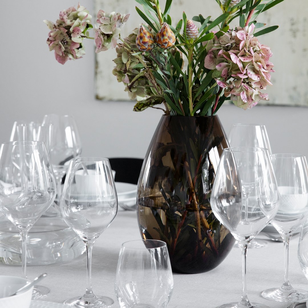 Elegant dark brown vase with floral arrangement, surrounded by clear glassware on a neutral table setting.