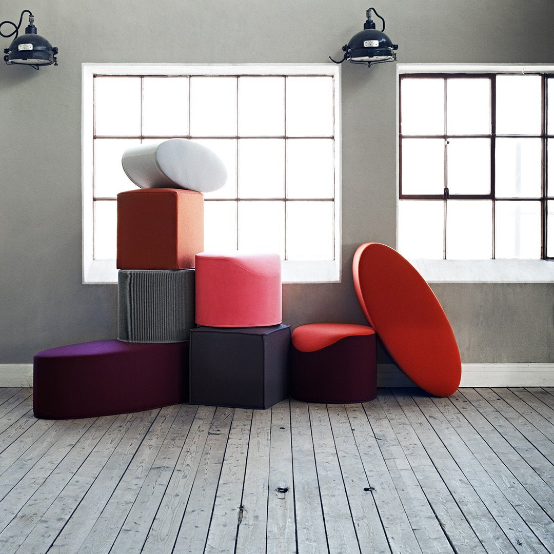 Colorful seating poufs in various shapes: red, pink, gray, orange, and purple, stacked against a light gray wall.