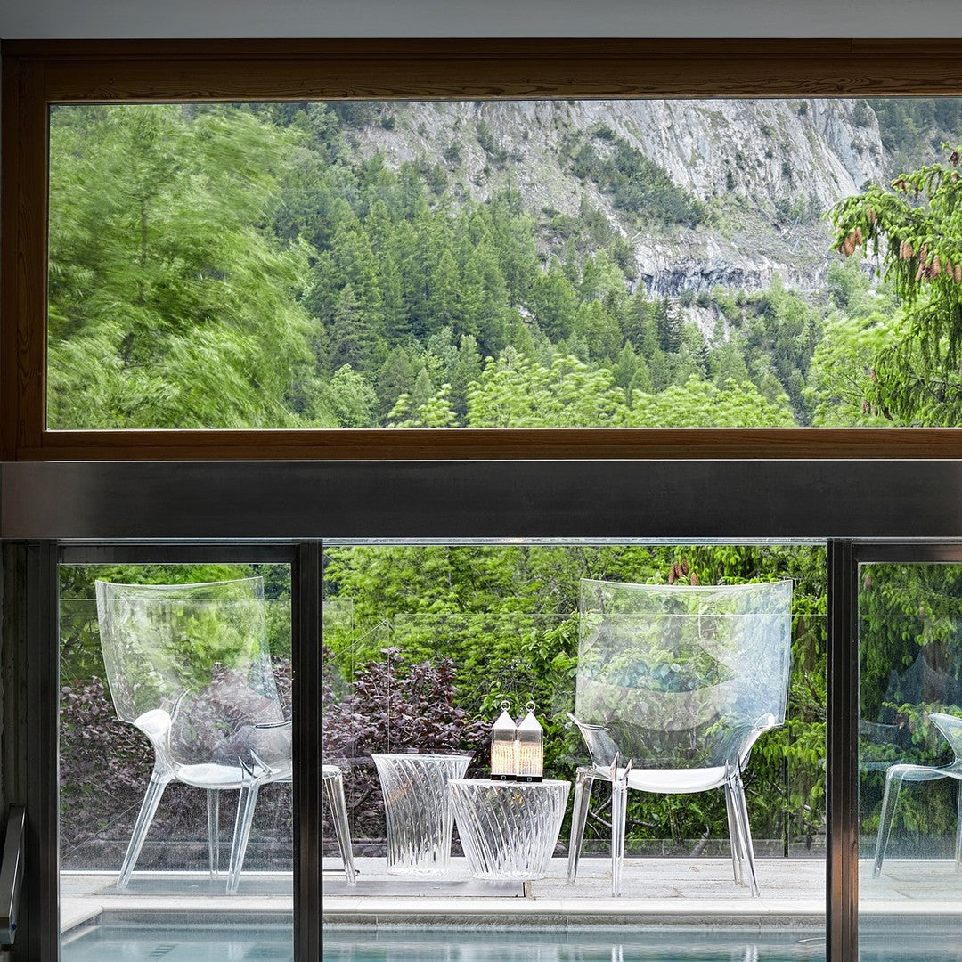 Clear chairs with a sleek design, glass table, and green natural backdrop. Modern and minimalist aesthetics.