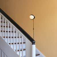 Modern black wall lamp with a circular white shade against a warm beige wall, complemented by a wooden staircase.