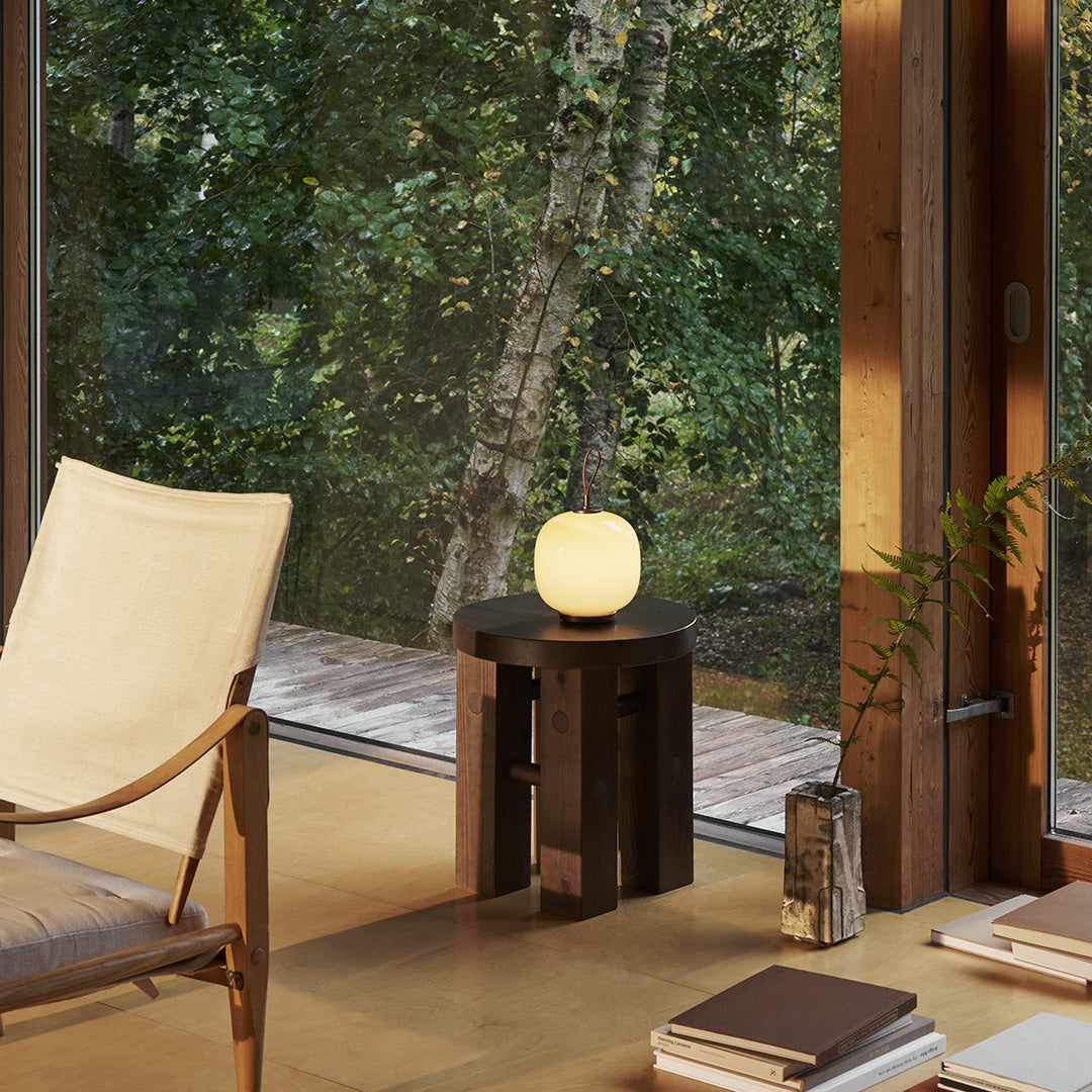 Soft white lamp on a dark wooden side table, surrounded by natural green foliage and warm neutral tones.
