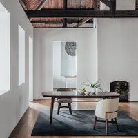 Modern dining table with a warm wood finish, paired with a light upholstery chair, set on a dark area rug.