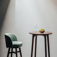 Green upholstered bar stool beside a round wooden table with a bowl of green grapes, set against a neutral backdrop.