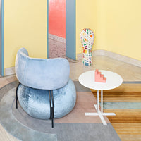 Light blue velvet chair with black legs, round white table, and colorful geometric sculpture against a pastel backdrop.