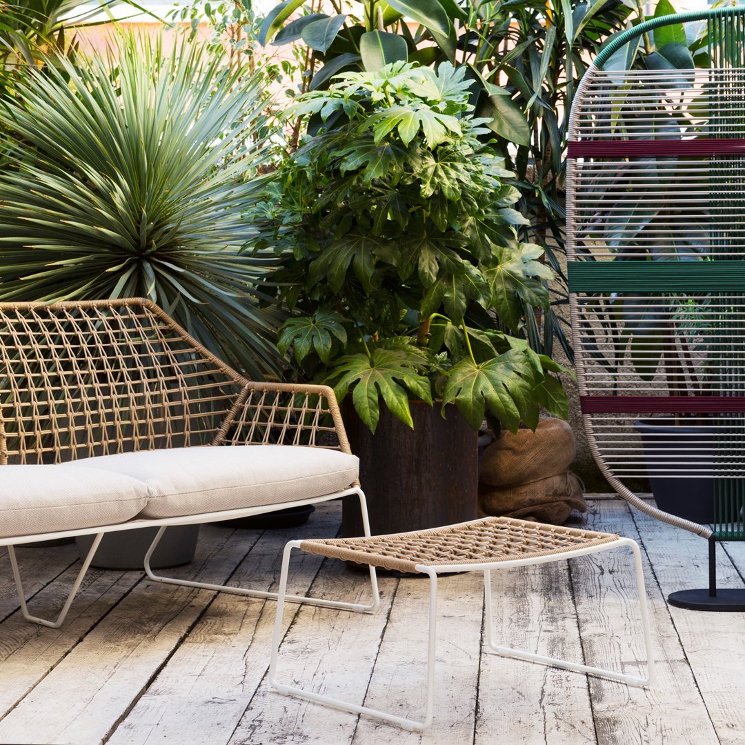 Light beige woven patio furniture with a geometric pattern, surrounded by lush green plants and colorful decor accents.