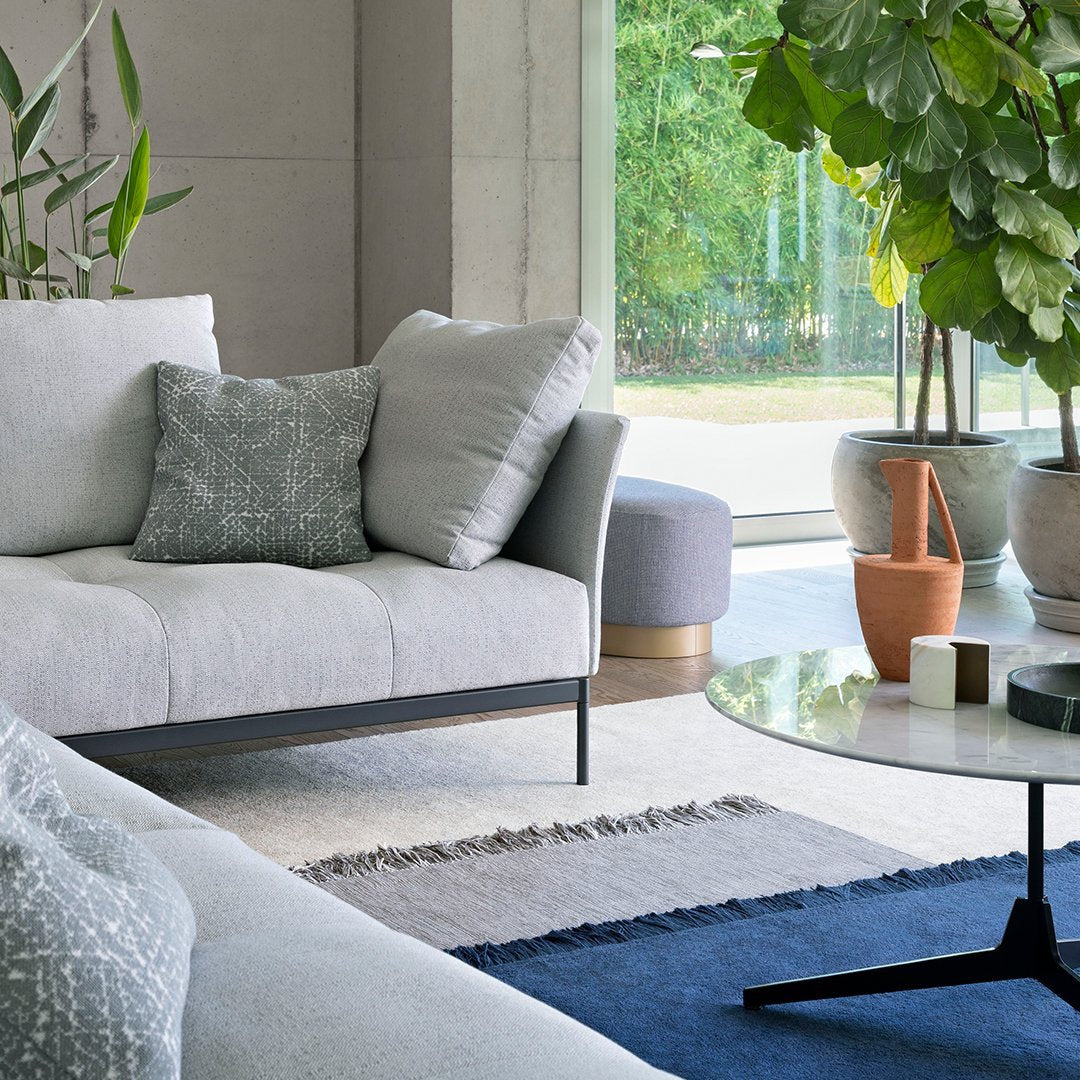 Light gray sofa with textured pillows, blue rug, and modern glass coffee table in a stylish living room setting.