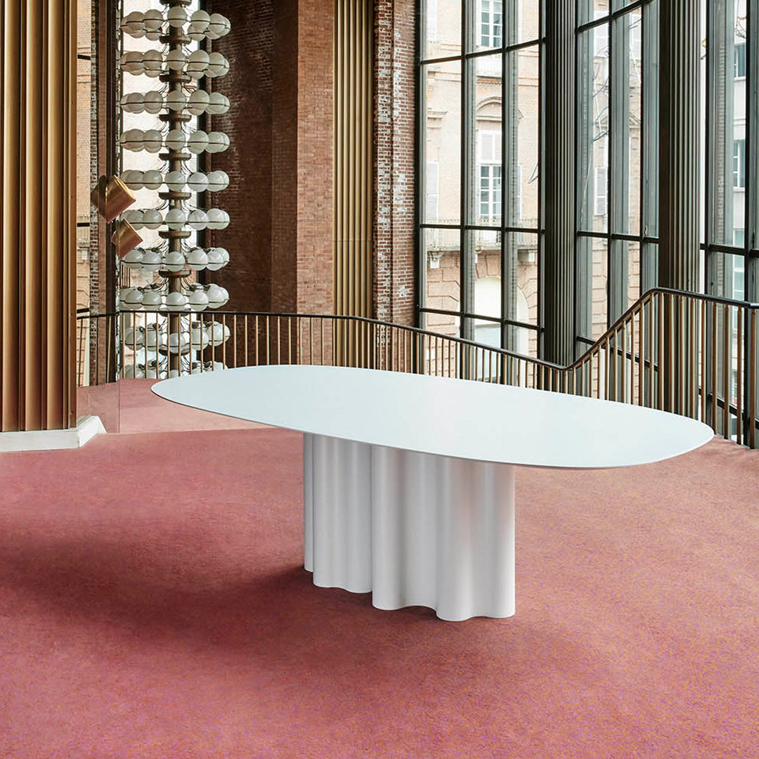 White oval table with fluted base on a pink carpet, set against large glass windows and textured brick walls.