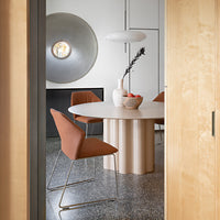 Modern beige dining table with circular top, paired with rust-colored chairs, set on a textured dark floor.