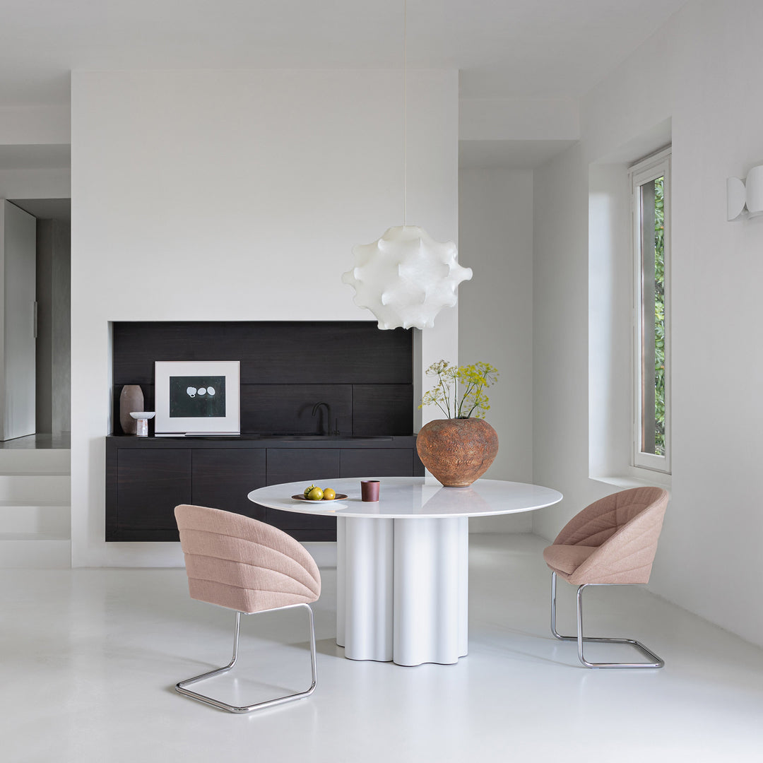 Modern dining area featuring a round white table, soft pink chairs, and a textured brown vase against a neutral backdrop.
