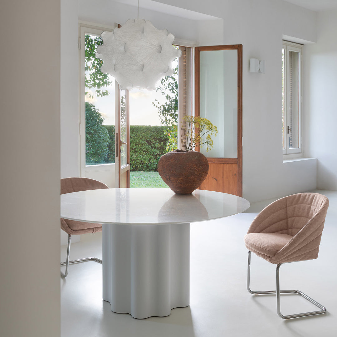 Modern round white table with a textured gray base, paired with soft pink chairs in a bright, minimalist room.