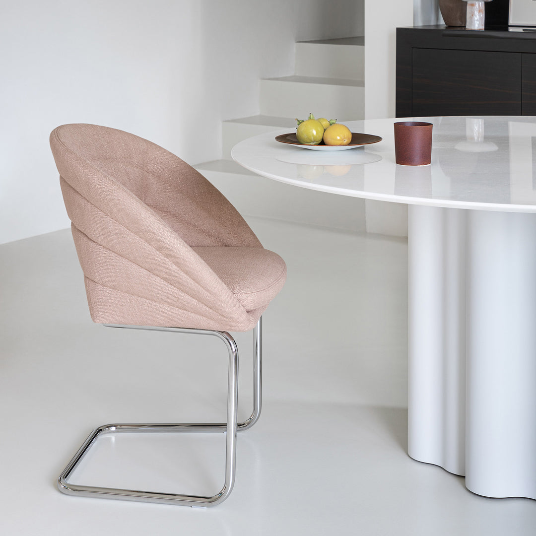 Pink upholstered chair with curved design against a modern white table and light floor. Simple and elegant aesthetic.