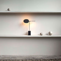 Black and gold modern lamp with a warm glow, displayed on a minimalist white shelf with gray stones.