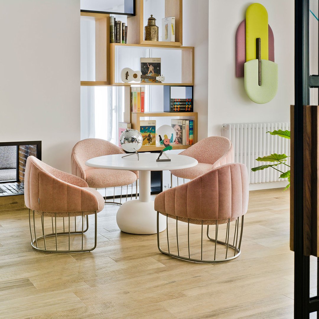 Blush pink upholstered chairs with sleek metallic bases around a white round table, set against a bright, modern interior.