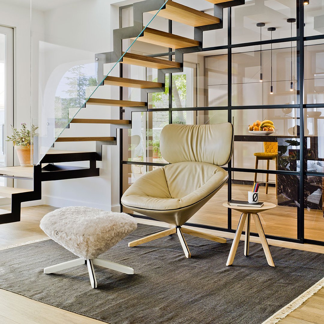Beige leather chair with a soft white ottoman on a dark gray rug, surrounded by elegant wood and glass accents.