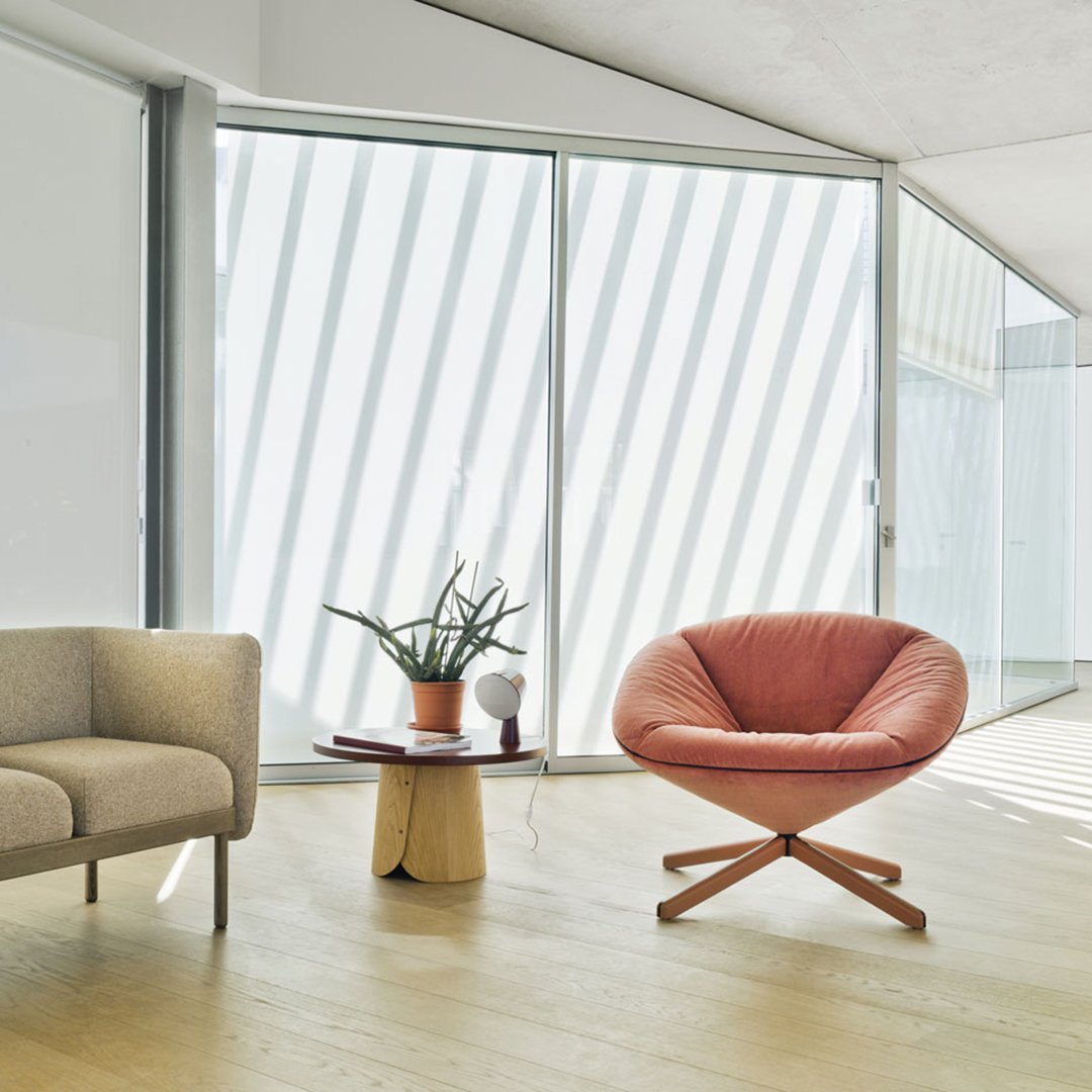 Peach curved chair with wood legs, accompanied by a beige armchair and light wood table in a sunlit, striped-patterned room.