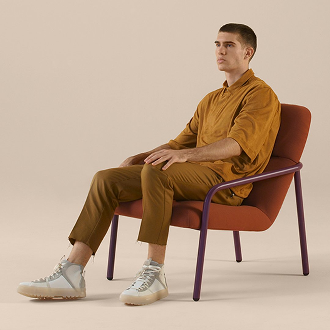 "Man in mustard shirt and pants sitting in a burgundy chair with purple legs, against a neutral background."