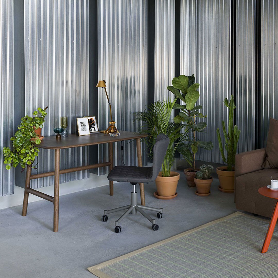 "Stylish gray desk chair on a light gray floor, surrounded by green plants and a patterned area rug in a modern space."
