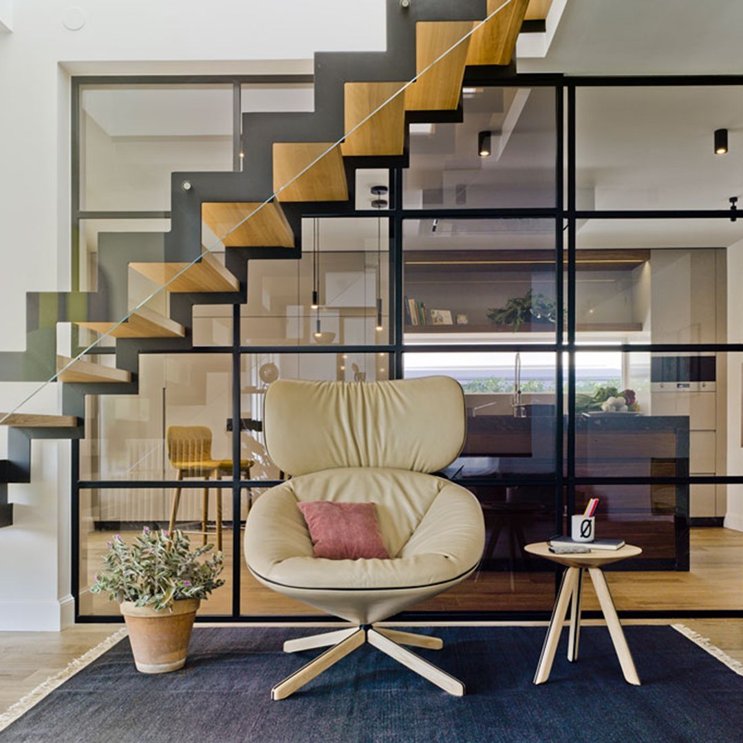 Beige armchair with a pink accent cushion on a dark blue rug, set against modern glass and wood staircase.